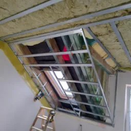 Poddasze w trakcie ocieplania z widoczną konstrukcją metalową pod płyty karton-gips, wełną mineralną, folią paroizolacyjną, oknem dachowym i drabiną z narzędziami.