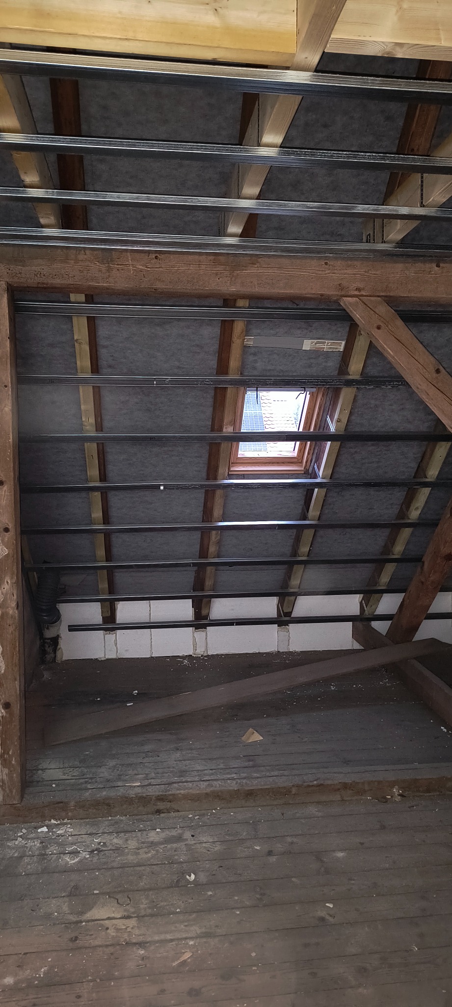 Poddasze w trakcie przygotowania do izolacji, widoczne drewniane krokwie, metalowy stelaż pod zabudowę, okno dachowe i częściowo ocieplone ściany.