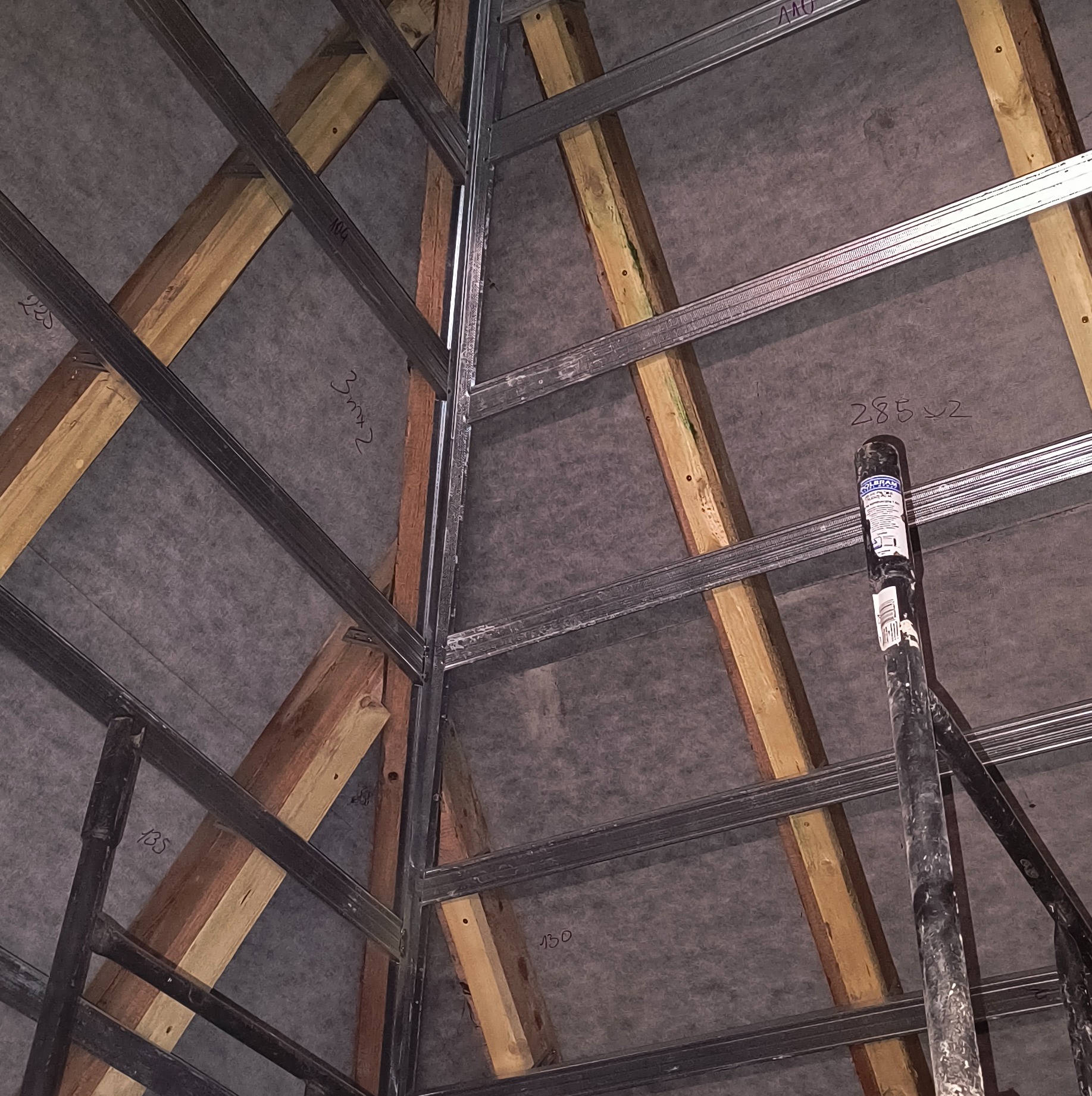 Konstrukcja metalowa i drewniana poddasza z widoczną szarą membraną, z naniesionymi odręcznymi numerami na membranie, wsparta na rusztowaniu.