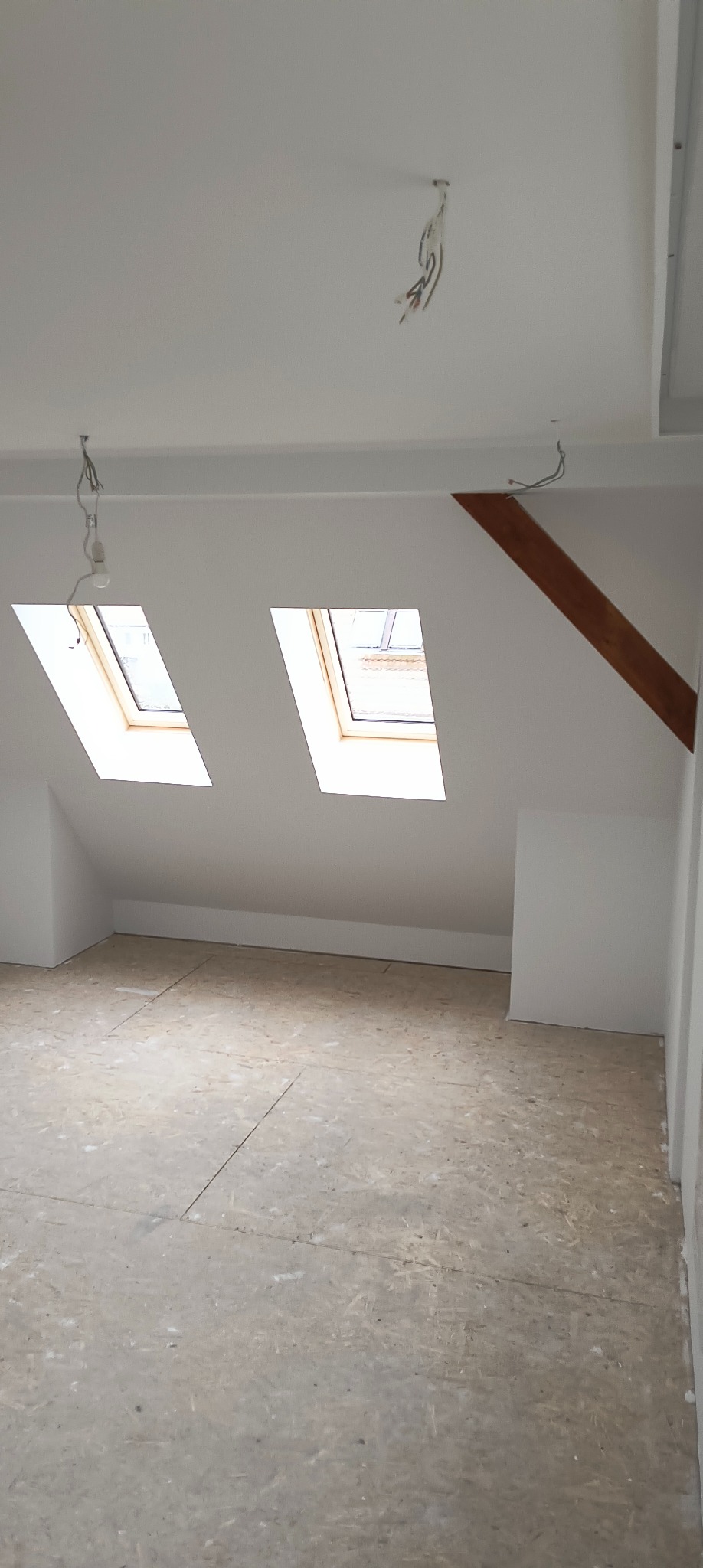 Poddasze z dwoma oknami dachowymi, surową podłogą z płyt OSB i odsłoniętymi przewodami elektrycznymi zwisającymi z sufitu.