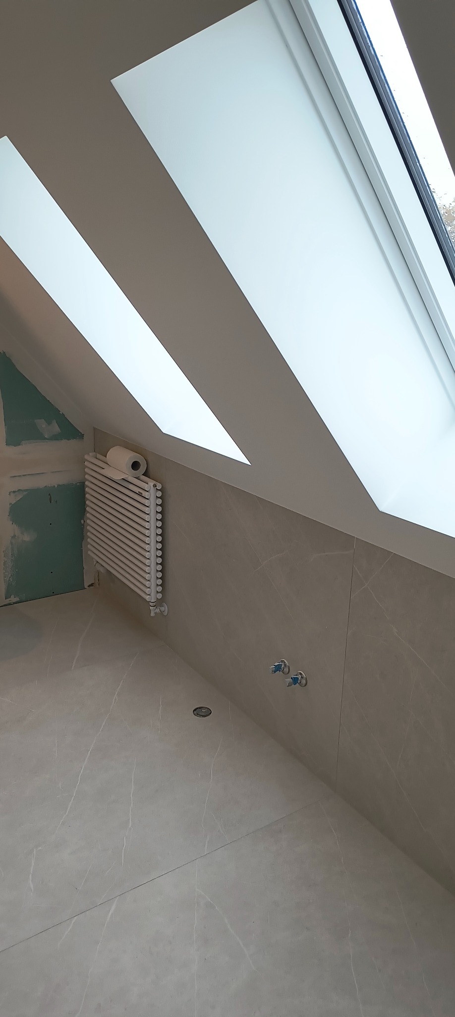 Poddasze ze skosami i oknami dachowymi w trakcie wykańczania, widoczne płytki imitujące marmur, grzejnik z rolką papieru toaletowego i przygotowane przyłącza wodne.