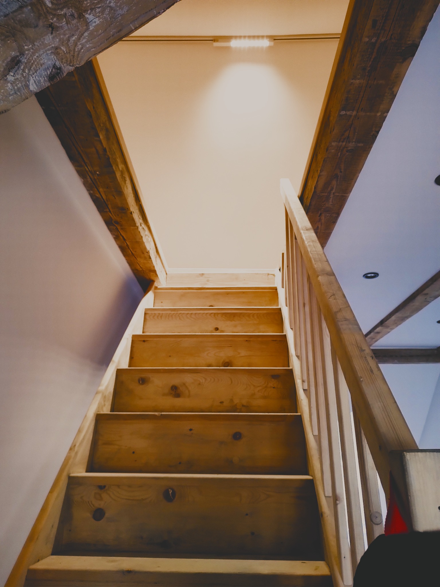 Drewniane schody z balustradą prowadzące na poddasze z widocznymi belkami stropowymi, oświetlone światłem punktowym.