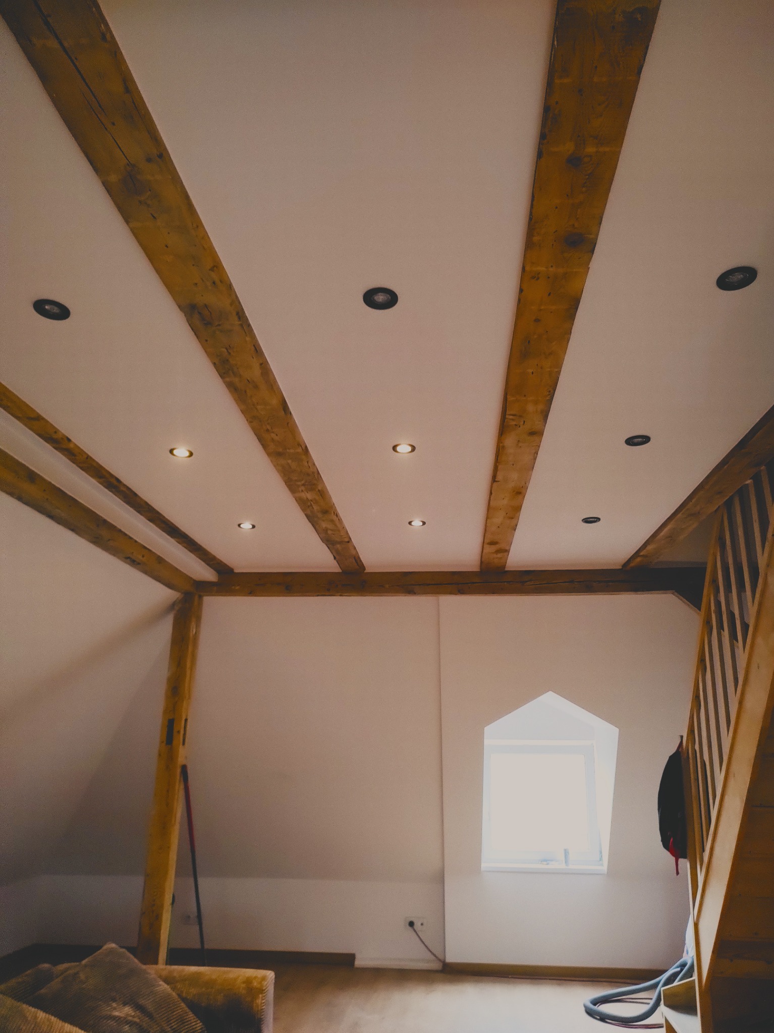 Poddasze z drewnianymi belkami stropowymi, oświetleniem punktowym i małym oknem w kształcie trapezu, widoczne fragmenty schodów i mebli.