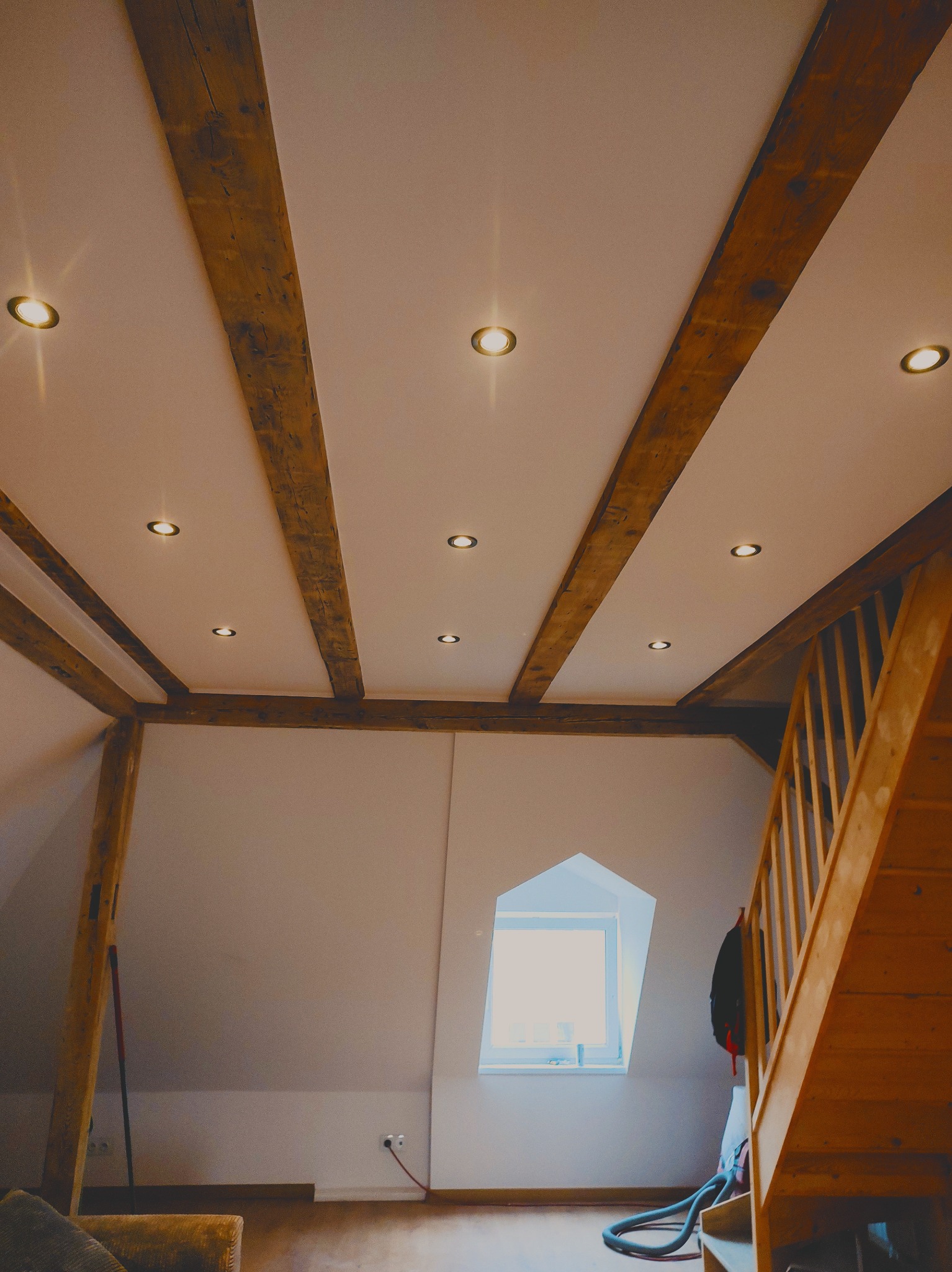 Poddasze po remoncie z drewnianymi belkami stropowymi, oświetleniem punktowym w suficie, drewnianymi schodami i oknem w szczycie ściany
