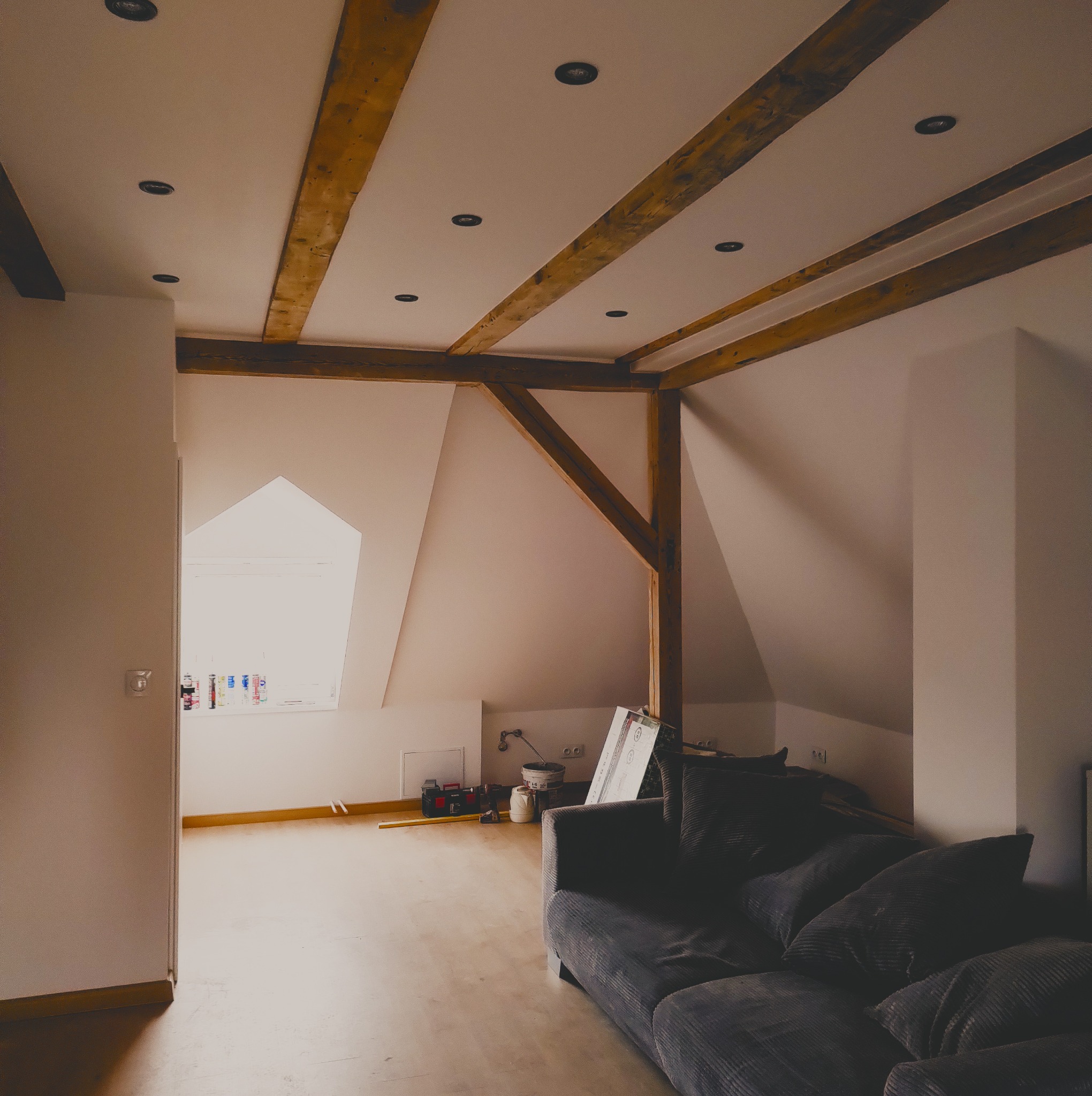 Poddasze z drewnianymi belkami stropowymi i oświetleniem punktowym, częściowo wykończone, z widocznym oknem dachowym, narzędziami i kanapą w tle.