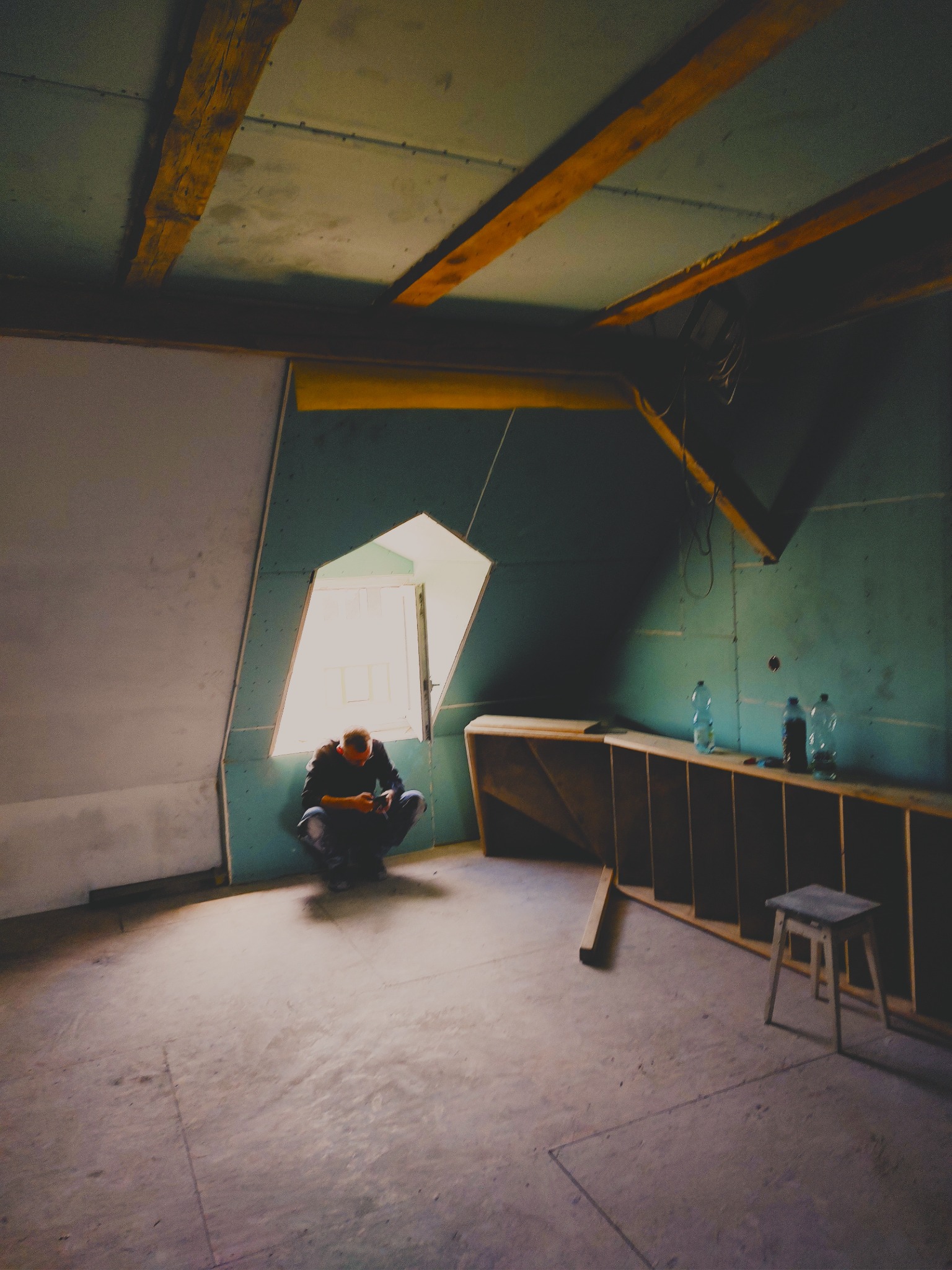 Adaptacja poddasza: mężczyzna siedzi w oknie w pokoju z drewnianymi belkami stropowymi, ścianami pokrytymi płytami i konstrukcją z drewna, butelki z wodą na półce.