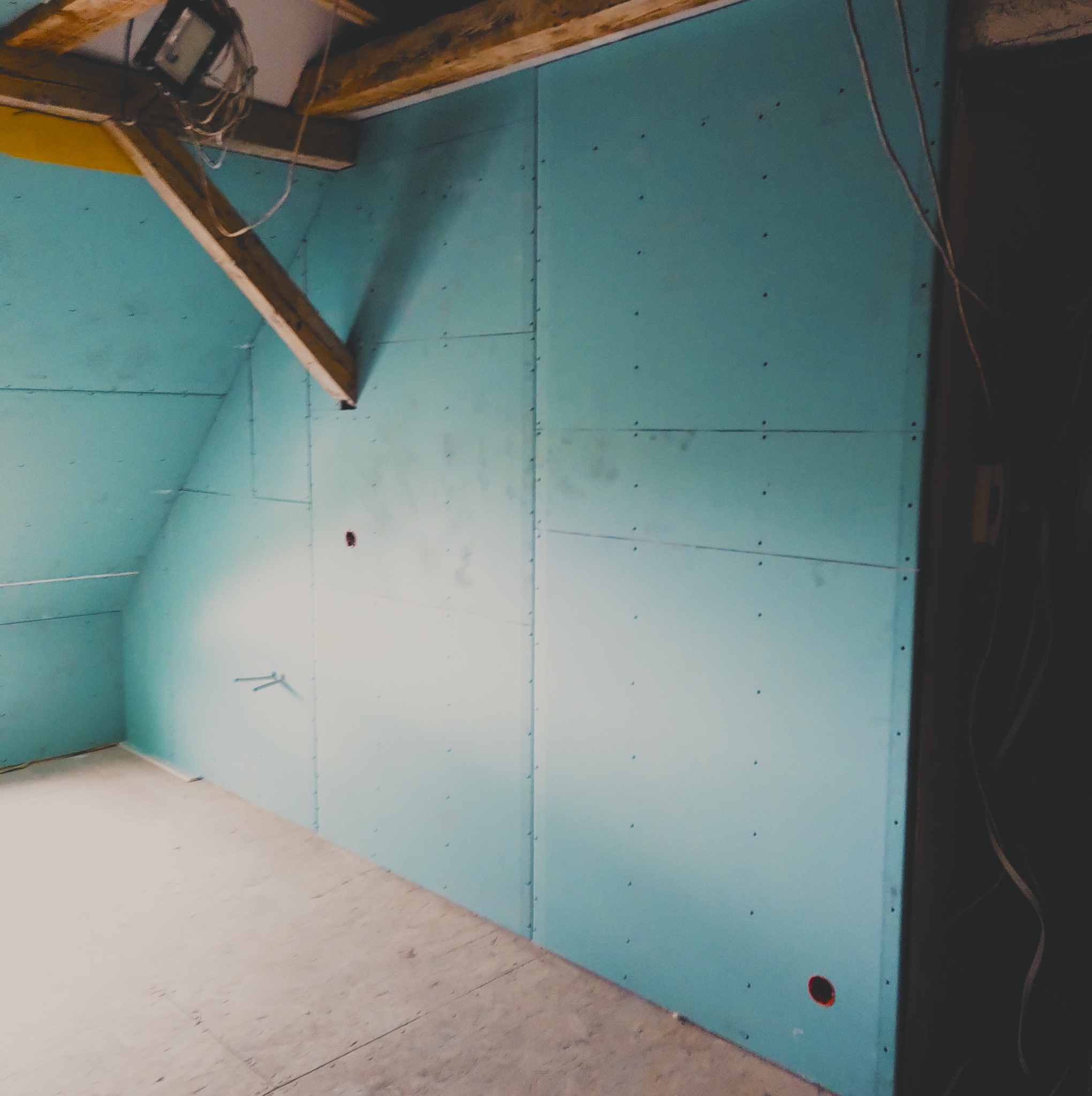 Poddasze z zamontowanymi niebieskimi płytami na ścianach, widoczne drewniane elementy konstrukcyjne i lampa halogenowa na suficie, oraz otwór na gniazdko elektryczne.