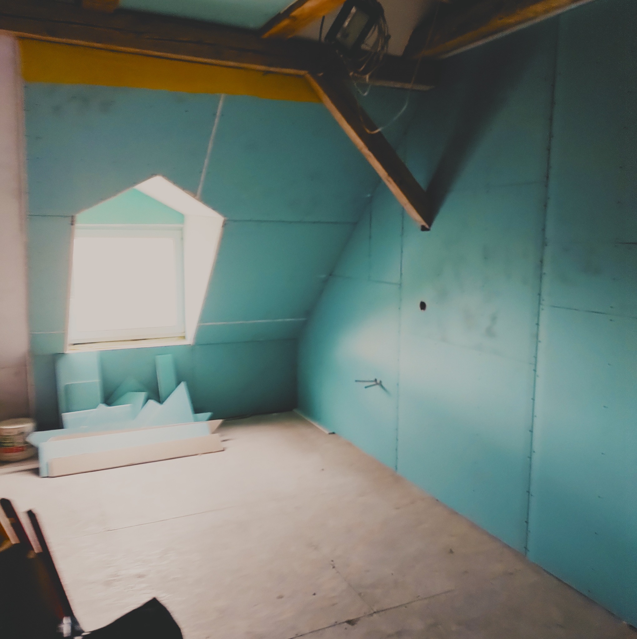 Poddasze z oknem dachowym, wykończone niebieską płytą gipsowo-kartonową. Widoczne drewniane elementy konstrukcyjne dachu, fragment podłogi i narzędzia w rogu.