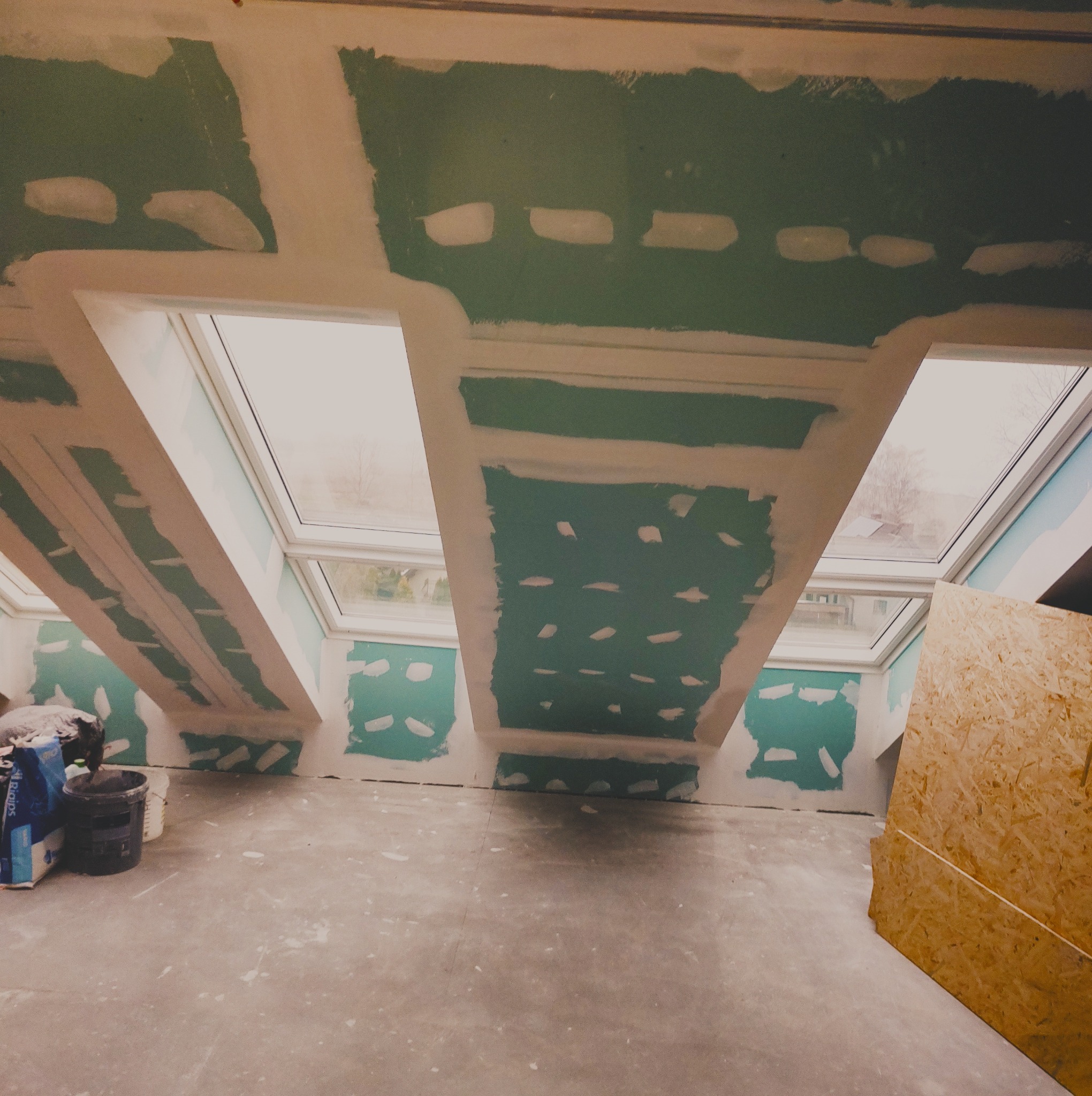 Poddasze w trakcie prac wykończeniowych: widoczne płyty na skosach i suficie, spoiny wypełnione masą, trzy okna dachowe w konstrukcji, surowe ściany i podłoga, narzędzia i materiały budowlane w tle.