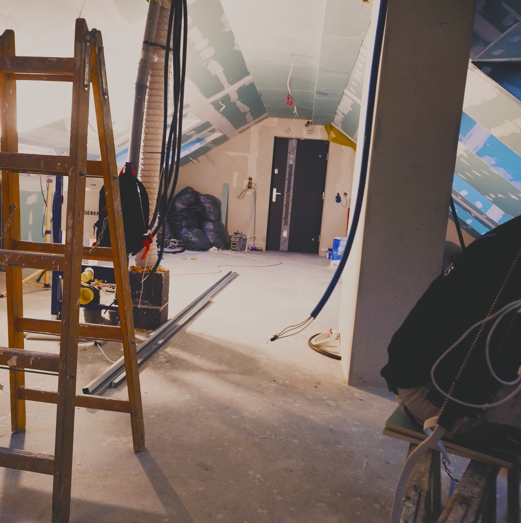 Poddasze w trakcie prac wykończeniowych: widoczne płyty gipsowo-kartonowe na skosach, metalowe profile na podłodze, stara drewniana drabina i elementy instalacji elektrycznej.