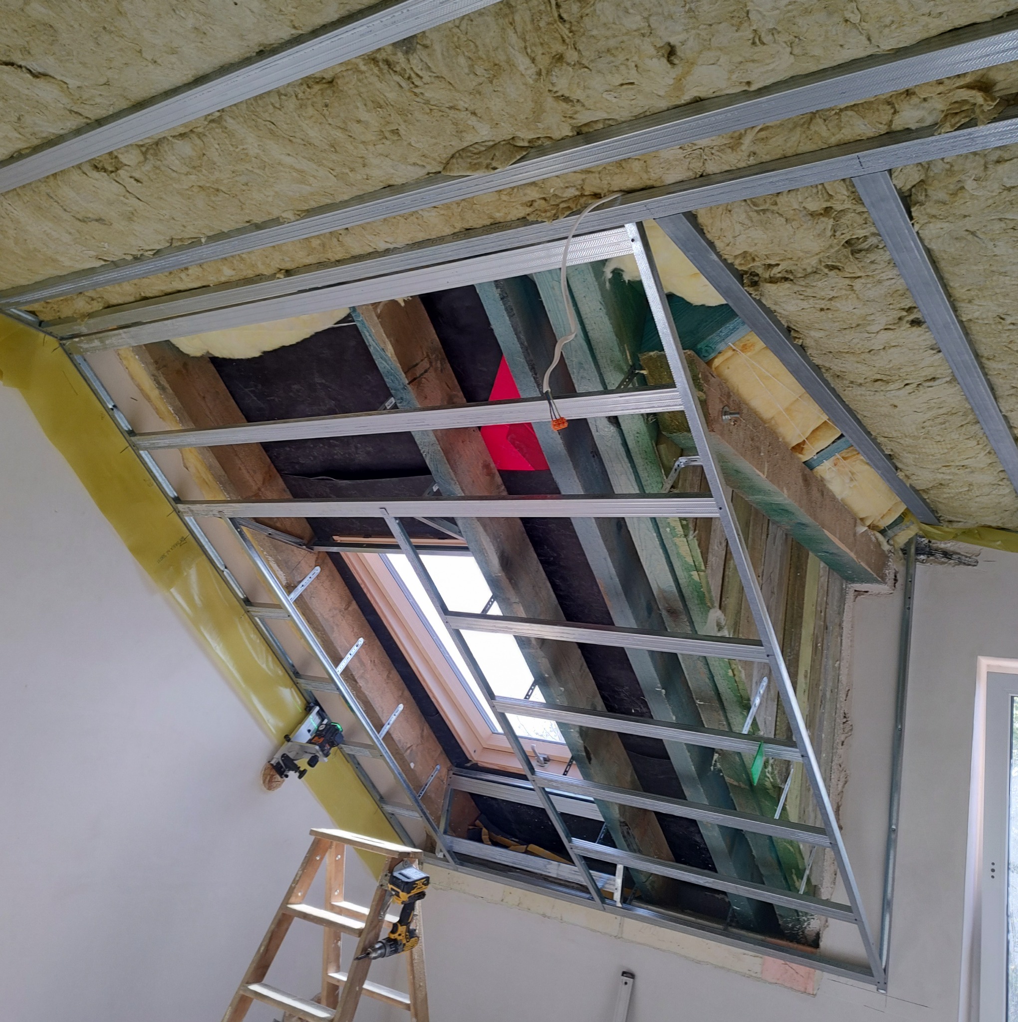 Poddasze w trakcie ocieplania z widoczną konstrukcją metalową pod płyty karton-gips, wełną mineralną, folią paroizolacyjną, oknem dachowym i drabiną z narzędziami.