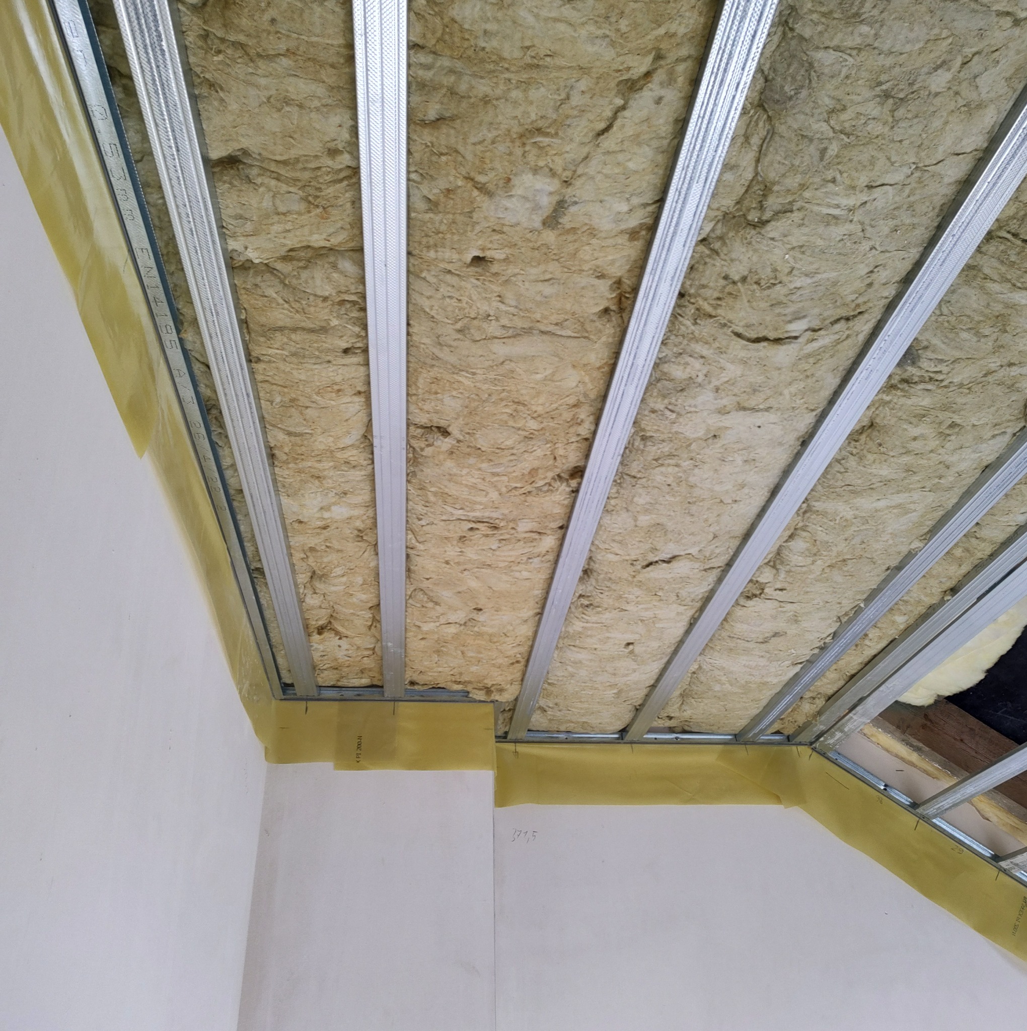 Sufit podwieszany w trakcie montażu: widoczne metalowe profile konstrukcyjne wypełnione wełną mineralną, zabezpieczone folią paroizolacyjną przy ścianach.