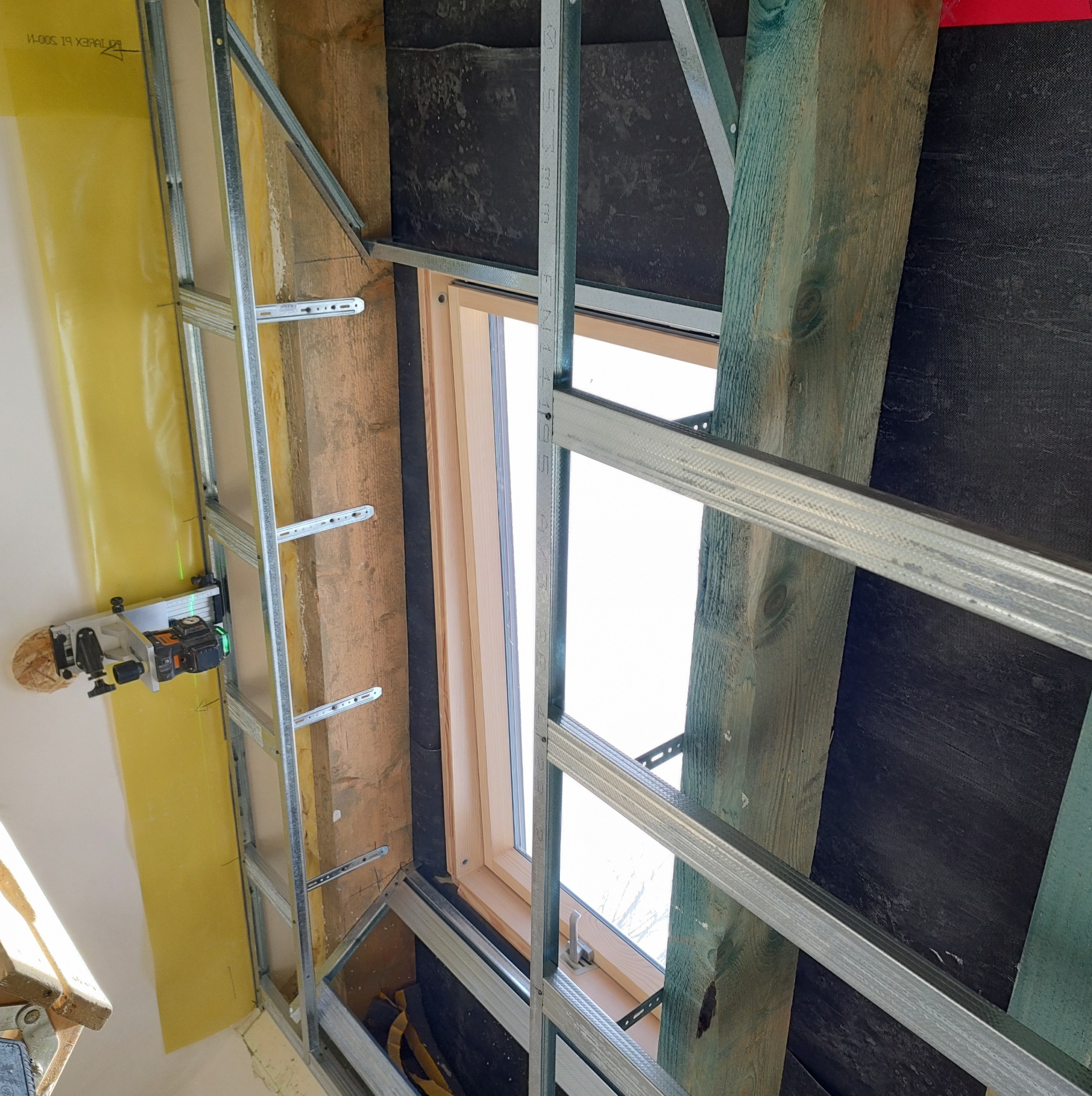 Montaż metalowego rusztu pod okno na poddaszu, widoczna żółta izolacja i drewniana konstrukcja dachu, użycie poziomicy laserowej.