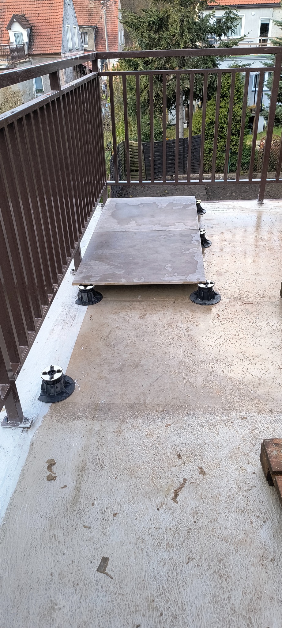 Montaż podłogi na regulowanych wspornikach na balkonie z brązową balustradą. Widoczne płyty podłogowe i elementy konstrukcyjne. Prace wykończeniowe na zewnątrz.