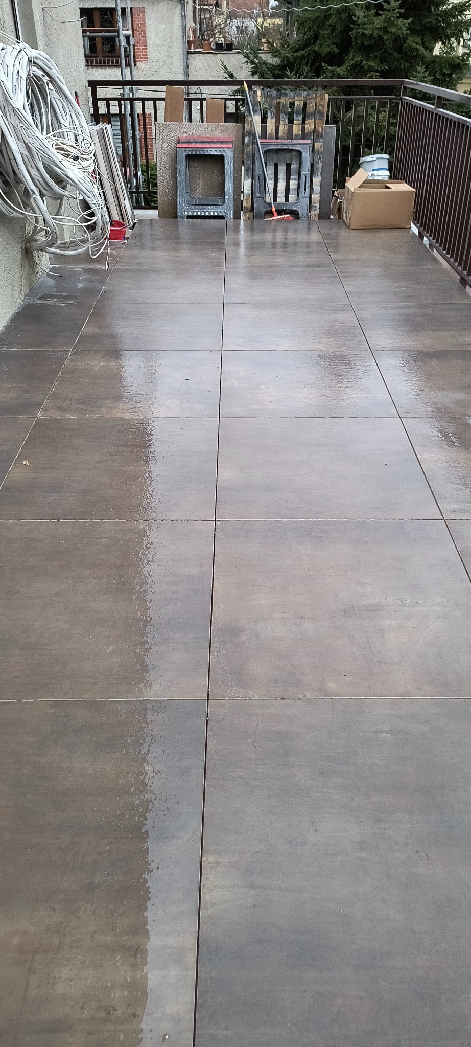 Wyłożony brązowymi płytkami taras po deszczu, widoczne narzędzia i materiały budowlane przy ścianie i balustradzie. Układanie płytek na zewnątrz.