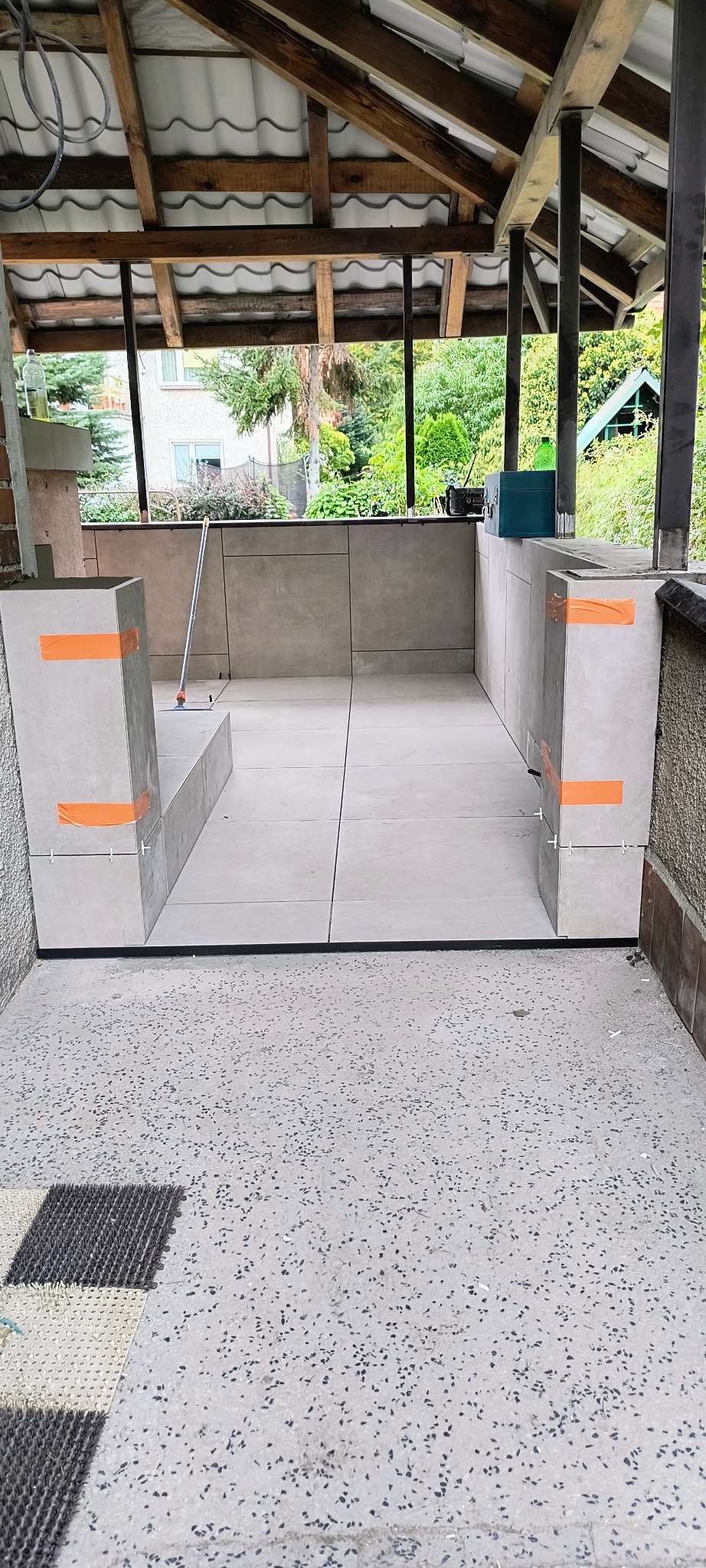 Wykończony taras z szarymi płytkami, zabezpieczony pomarańczową taśmą. Widoczne fugi i elementy konstrukcyjne z drewna. Tło z zielenią drzew.