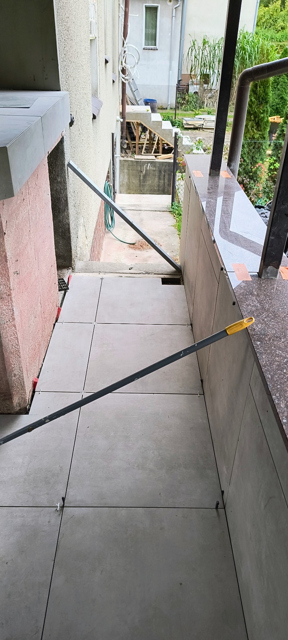 Układanie szarych płytek na zewnątrz budynku, widoczne krzyżyki dystansowe i podpory. Prace wykończeniowe na schodach zewnętrznych.