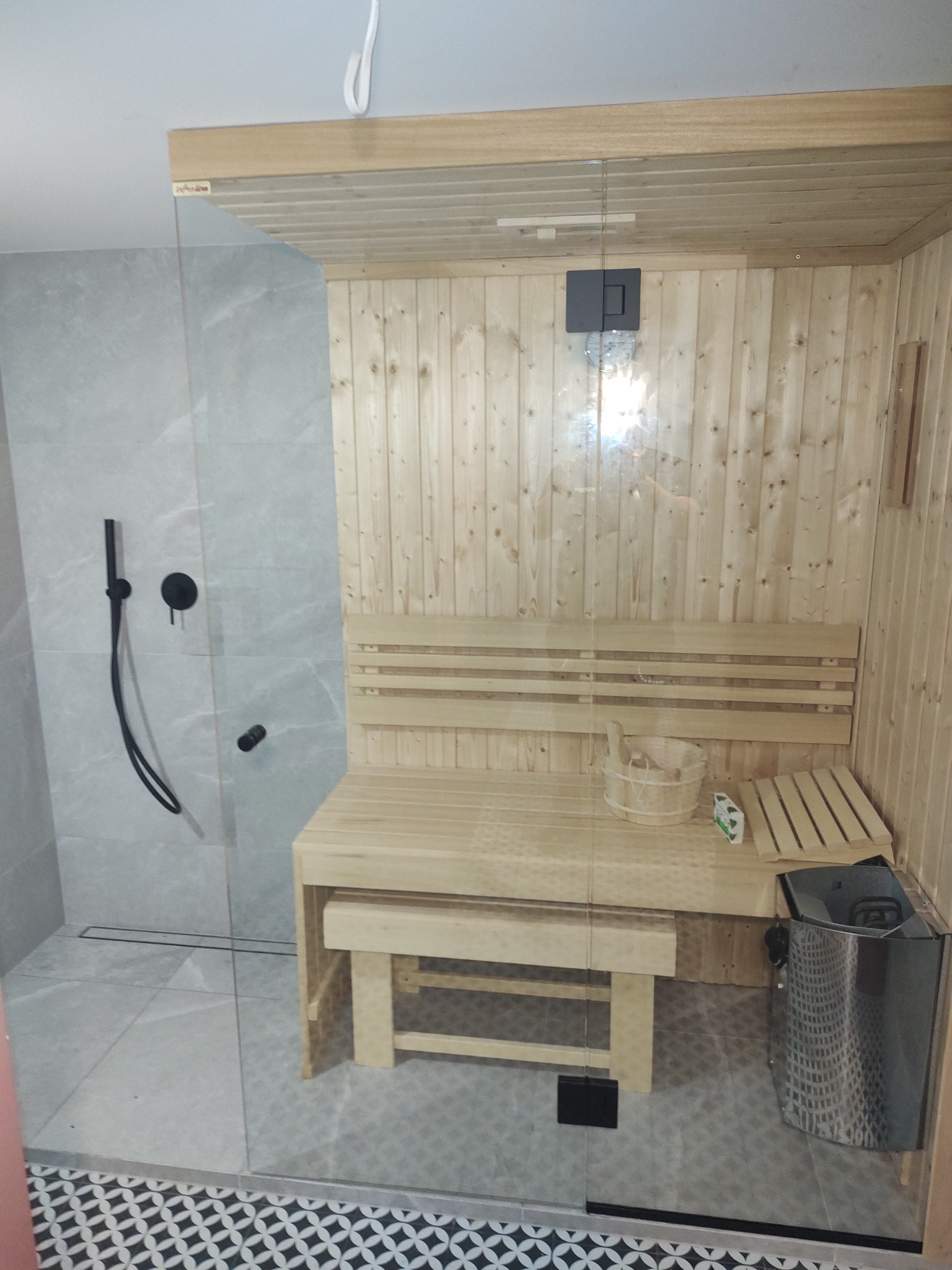 Nowoczesna sauna w łazience: drewniane wykończenie, szklana kabina prysznicowa z czarną armaturą, szare płytki i dekoracyjna podłoga.