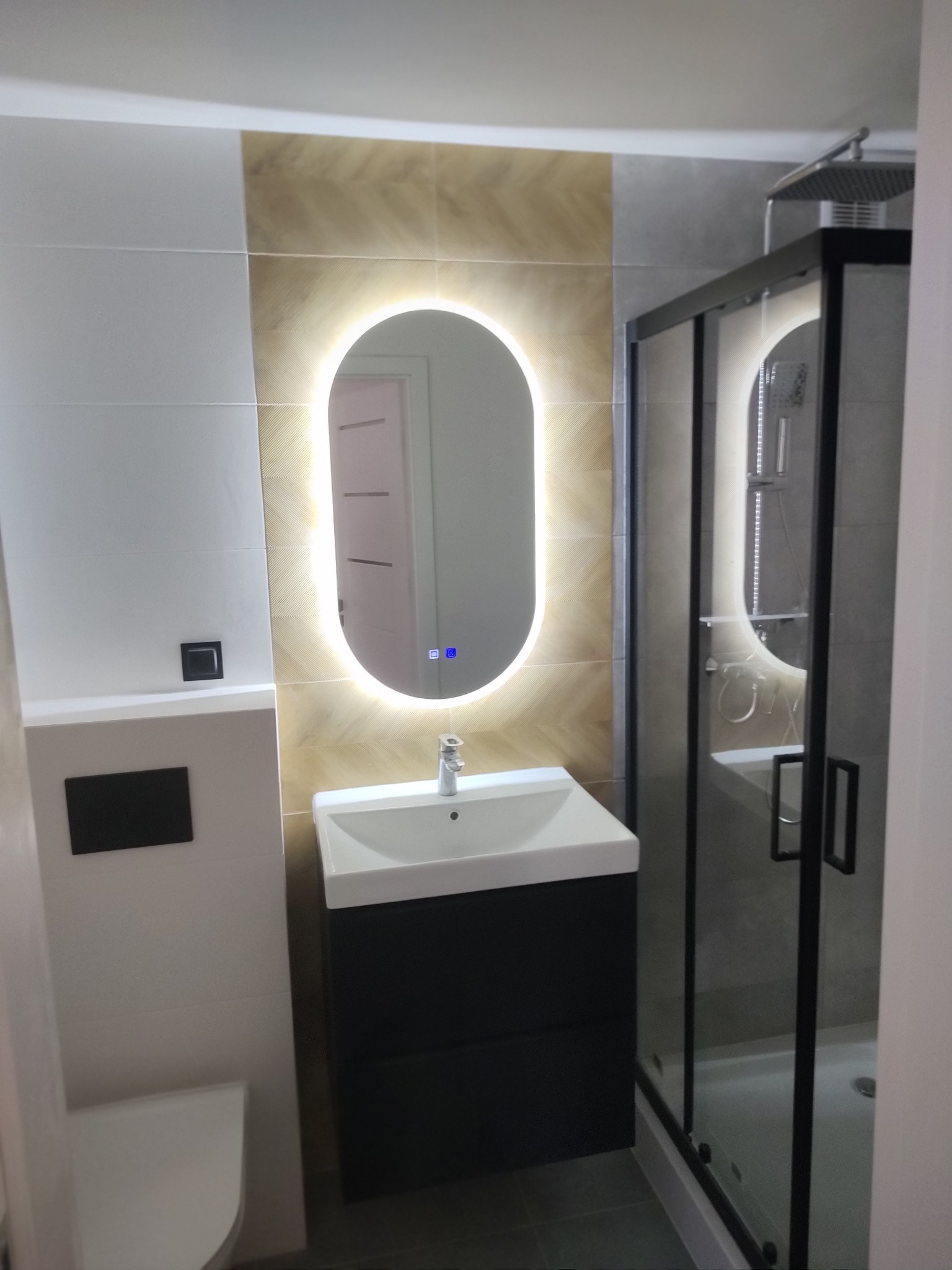 Nowoczesna łazienka po remoncie: owalne lustro z podświetleniem LED, czarna szafka z umywalką, kabina prysznicowa z czarnymi ramami i płytki w odcieniach szarości i beżu.