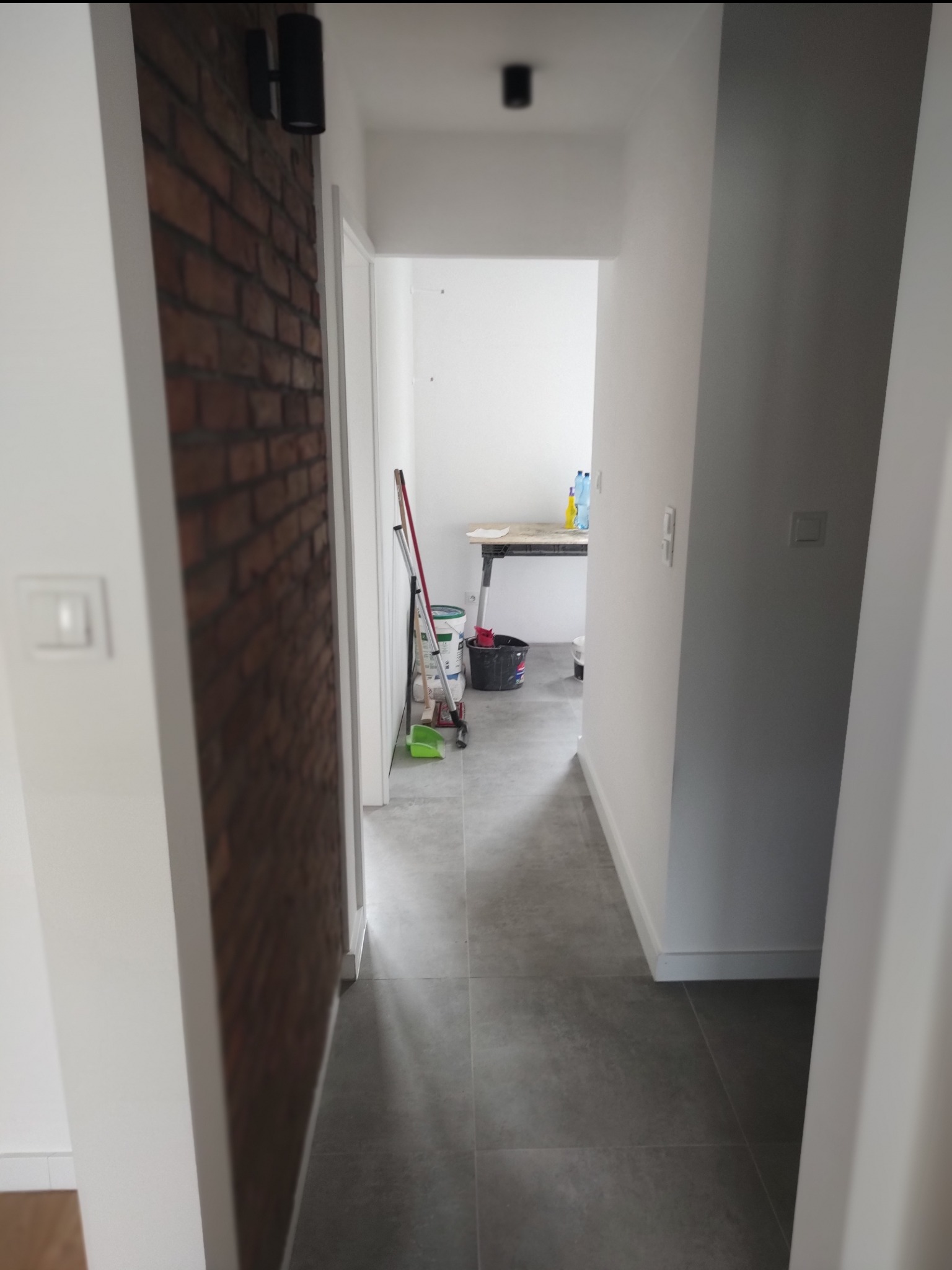 Korytarz po remoncie z szarą podłogą i ścianą z cegły. Widoczne narzędzia malarskie i wiadro. Oświetlenie sufitowe.
