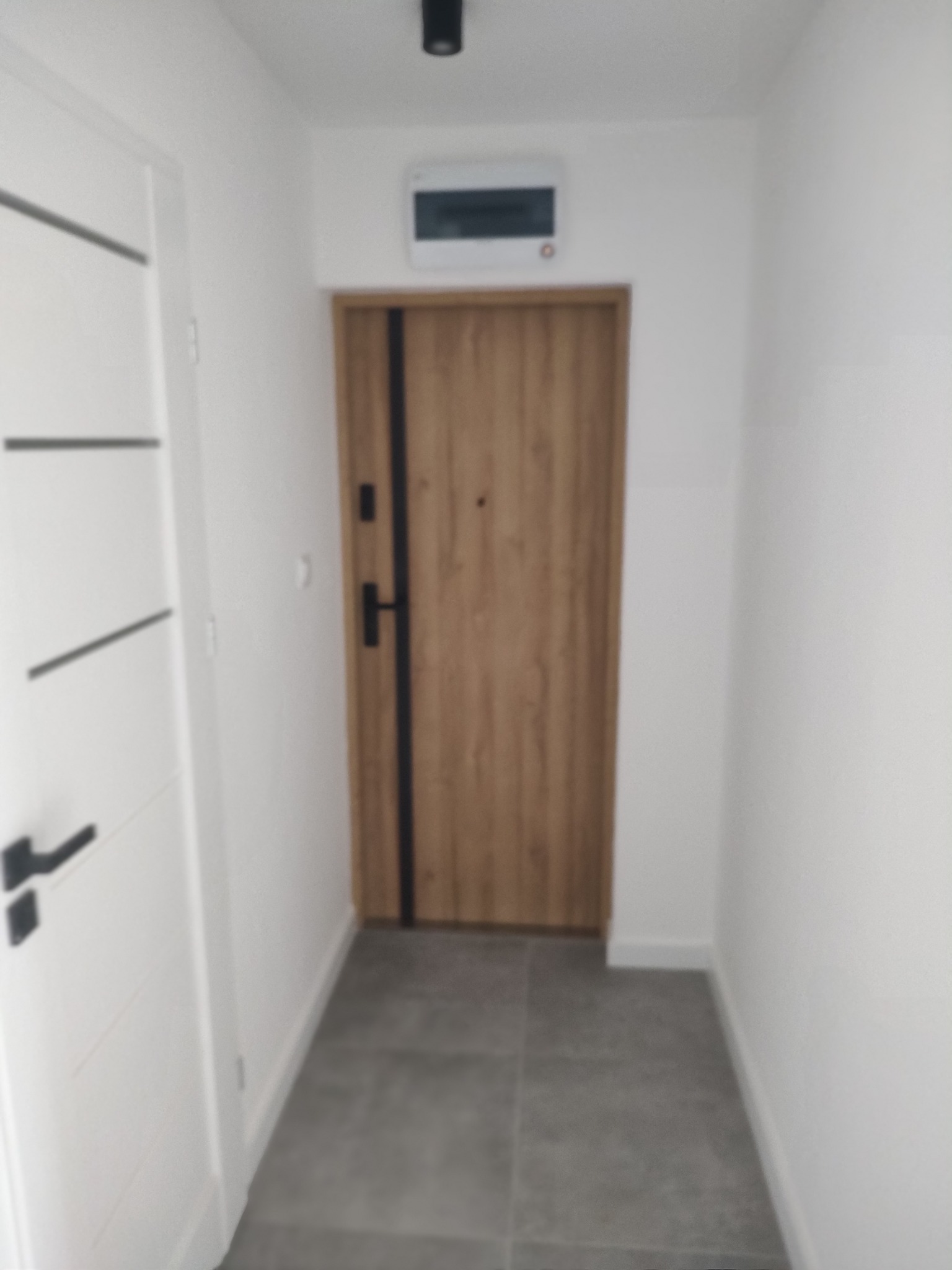 Nowoczesny przedpokój po remoncie: białe ściany, szara podłoga, drewniane drzwi z czarnymi akcentami i białe drzwi wewnętrzne z poziomymi frezami.