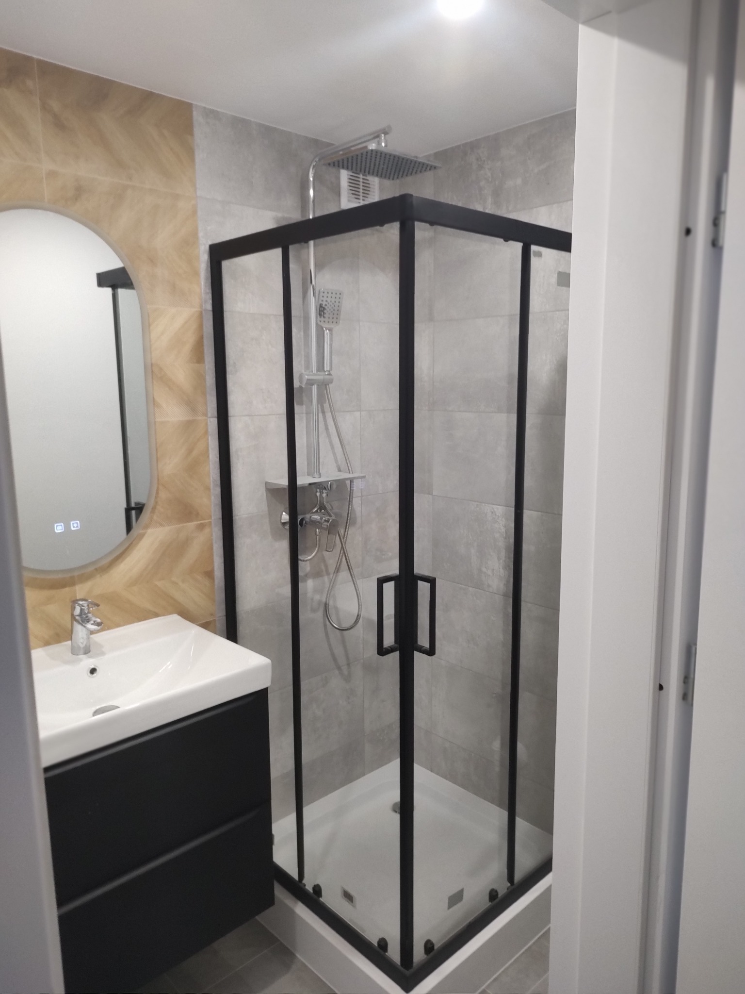 Nowoczesna łazienka po remoncie: kabina prysznicowa z czarną ramą, umywalka z szafką w kolorze antracytowym, owalne lustro z podświetleniem LED i płytki w odcieniach szarości i beżu.