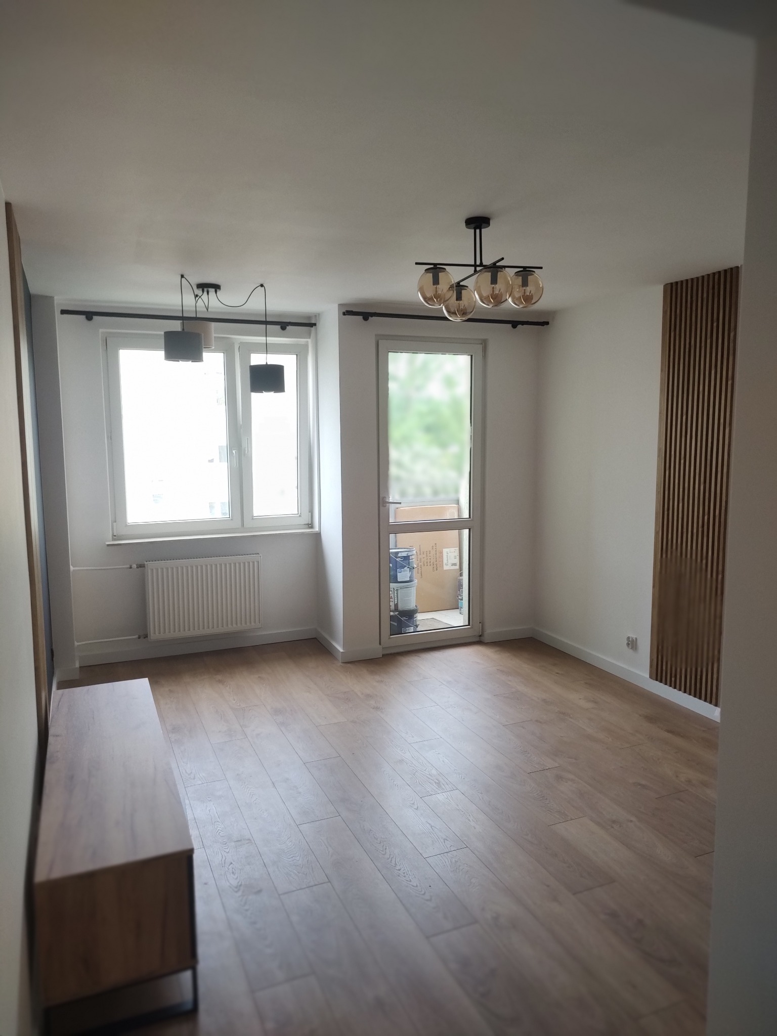 Jasne wnętrze po remoncie: drewniana podłoga, dekoracyjne panele ścienne, nowoczesne lampy sufitowe. Widok na okno i drzwi balkonowe. Minimalistyczny styl.