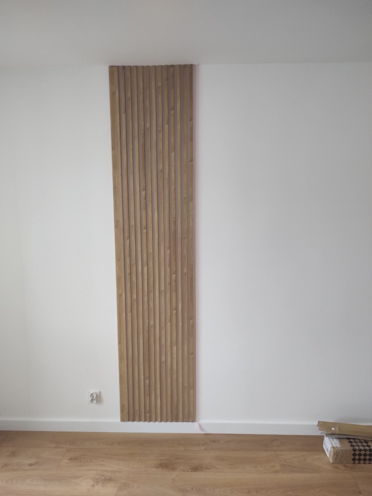 Drewniane lamele ścienne w pionowym układzie, zamontowane na białej ścianie. Widoczny fragment podłogi z paneli i gniazdko elektryczne. Minimalistyczny design wnętrza.