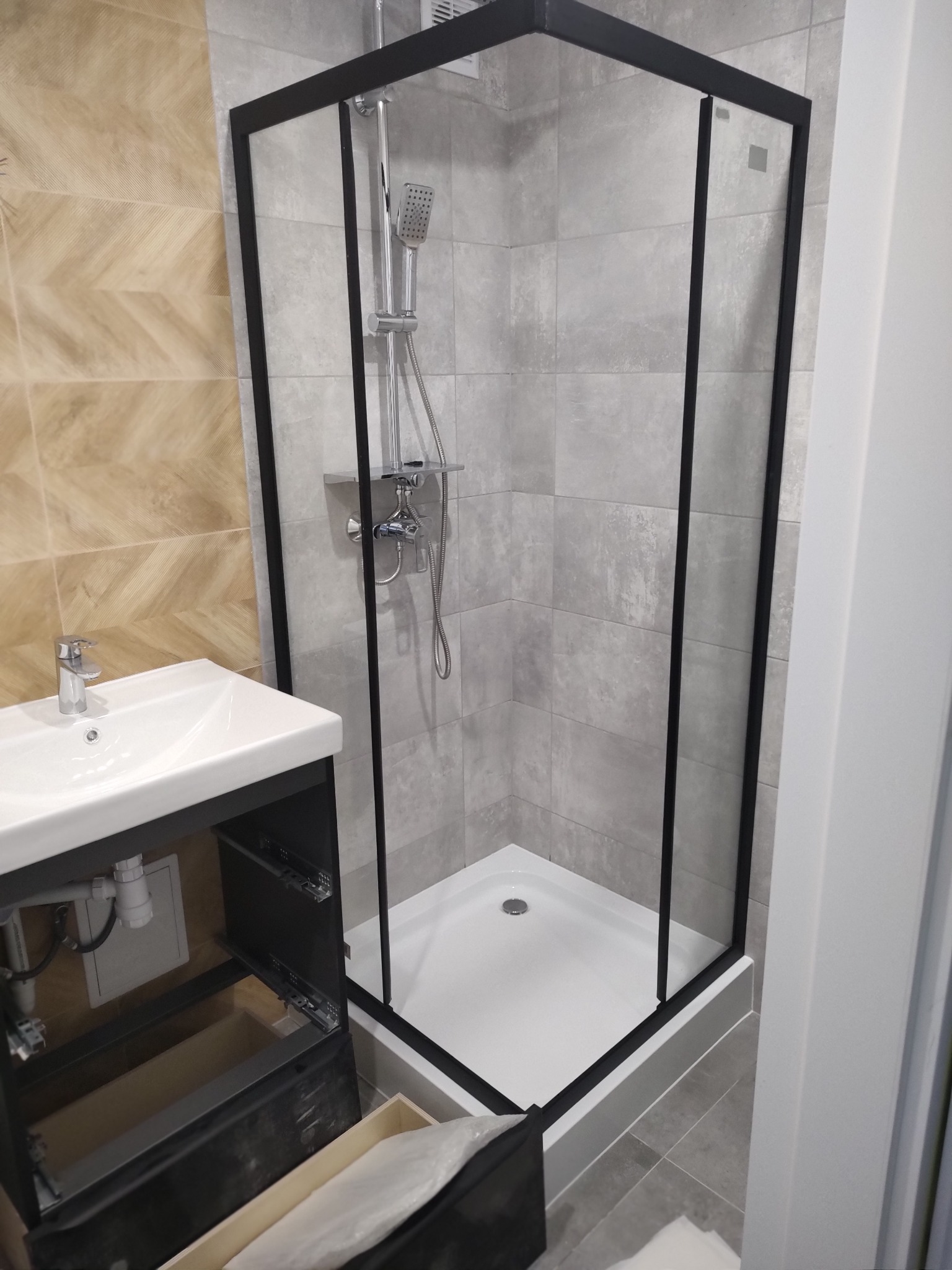 Nowoczesna kabina prysznicowa z czarną ramą i białą brodzikiem, umywalka z szafką w łazience wykończonej płytkami w odcieniach szarości i beżu.