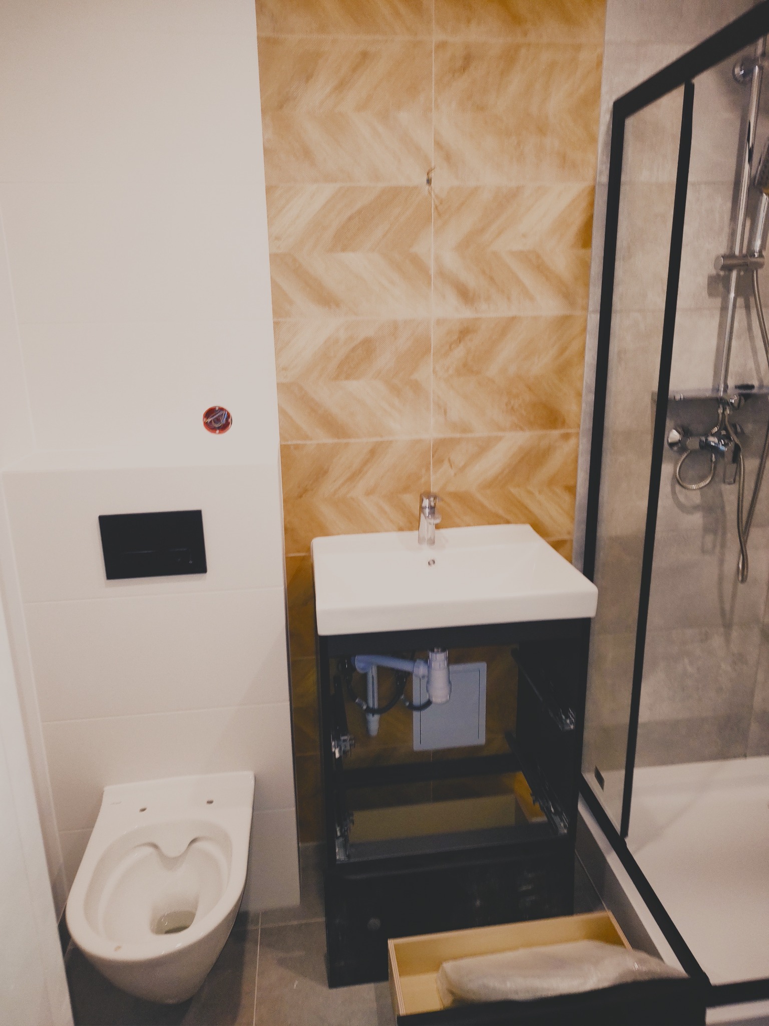 Nowoczesna łazienka po remoncie: podwieszana toaleta, umywalka z szafką (otwarte drzwiczki), kabina prysznicowa z czarną ramą, płytki imitujące drewno na ścianie.