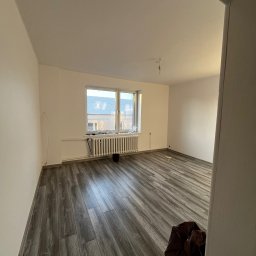 BRICHER Bartosz Brymora - Puste, jasne wnętrze po remoncie: świeżo pomalowane ściany, ułożone panele podłogowe w odcieniach szarości, grzejnik pod oknem. Widoczny worek z odpadami.