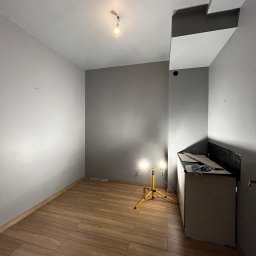 BRICHER Bartosz Brymora - Szare wnętrze po remoncie: drewnopodobne płytki na podłodze, świeżo pomalowane ściany w odcieniach szarości, żółty statyw z lampami oświetlającymi przestrzeń.