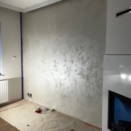 BRICHER Bartosz Brymora - Ściana z efektem dekoracyjnym w odcieniach szarości, zabezpieczona podłoga, widoczne narzędzia malarskie i puszka farby. Fragment kominka z boku kadru.