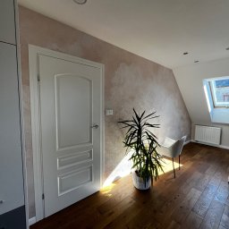 BRICHER Bartosz Brymora - Jasne wnętrze poddasza z drewnianą podłogą, ścianą w odcieniach beżu i białymi drzwiami. W rogu stoi roślina doniczkowa, a przez okno wpada naturalne światło.