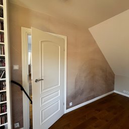 BRICHER Bartosz Brymora - Pokój na poddaszu z drewnianą podłogą i ścianą z dekoracyjną, beżową fakturą. Otwarta biała, panelowa  drzwi. Widoczny fragment regału z książkami.