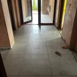 BRICHER Bartosz Brymora - Ułożona jasnoszara podłoga z płytek w korytarzu z otwartymi drzwiami na zewnątrz. Widoczne narzędzia glazurnicze i surowe ściany z fakturą.