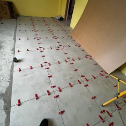 BRICHER Bartosz Brymora - Układanie szarych płytek podłogowych z czerwonymi klipsami poziomującymi. Widoczny fragment buta i lampa oświetleniowa. Żółta ściana w tle.