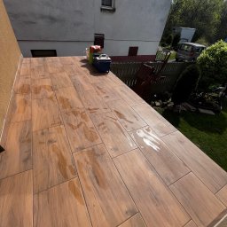 BRICHER Bartosz Brymora - Balkon wyłożony płytkami imitującymi drewno, z widocznymi śladami wody, sugerujący świeżo zakończone prace glazurnicze. W tle budynek i zieleń.