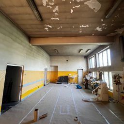 BRICHER Bartosz Brymora - Przygotowanie do prac w dużej sali: zabezpieczona podłoga, ściany z żółtym pasem, widoczne odrapania na suficie. W tle pracownik przy maszynie.