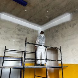 BRICHER Bartosz Brymora - Pracownik w kombinezonie maluje natryskowo sufit i ściany z rusztowania. Widoczne zabezpieczenia folią i taśmą malarską. Ściany w odcieniach szarości, sufit częściowo biały.