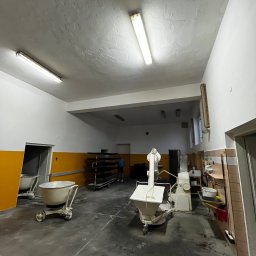 BRICHER Bartosz Brymora - Przestronne wnętrze piekarni z maszynami do ciasta, wózkiem na kółkach i regałem na blachy. Ściany biało-żółte, sufit z lampami. Widoczny fragment osoby w tle.