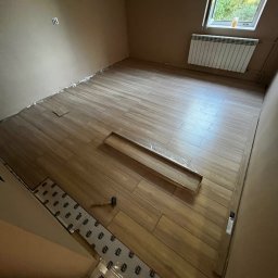 BRICHER Bartosz Brymora - Układanie paneli podłogowych w pokoju z beżowymi ścianami. Częściowo ułożone panele, widoczna folia izolacyjna i młotek. Okno z grzejnikiem w tle.