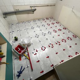 BRICHER Bartosz Brymora - Układanie płytek podłogowych w łazience z użyciem systemu poziomowania. Widoczne narzędzia glazurnicze i fragment osoby w obuwiu roboczym. Szare płytki, czerwone i niebieskie kliny.