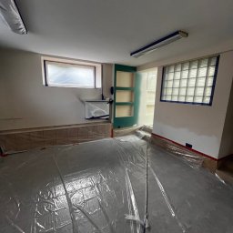 BRICHER Bartosz Brymora - Przygotowane pomieszczenie do malowania: podłoga zabezpieczona folią, okna i listwy oklejone taśmą malarską. Widoczne półki wnękowe i grzejnik.