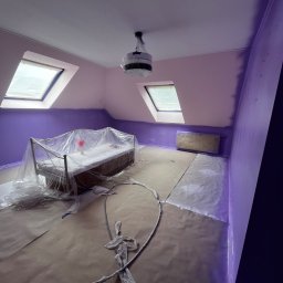 BRICHER Bartosz Brymora - Pokój na poddaszu w trakcie malowania na fioletowo. Meble zabezpieczone folią, widoczne skosy i okna dachowe. Częściowo pomalowane ściany.