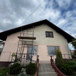 BRICHER Bartosz Brymora - Dom jednorodzinny w trakcie malowania elewacji. Widoczne rusztowanie, zabezpieczone okna folią i częściowo pomalowana ściana na jasny różowy kolor.