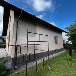 BRICHER Bartosz Brymora - Dom jednorodzinny w trakcie malowania elewacji, widoczne rusztowanie na tle zielonego trawnika i błękitnego nieba. Okno zabezpieczone folią malarską.