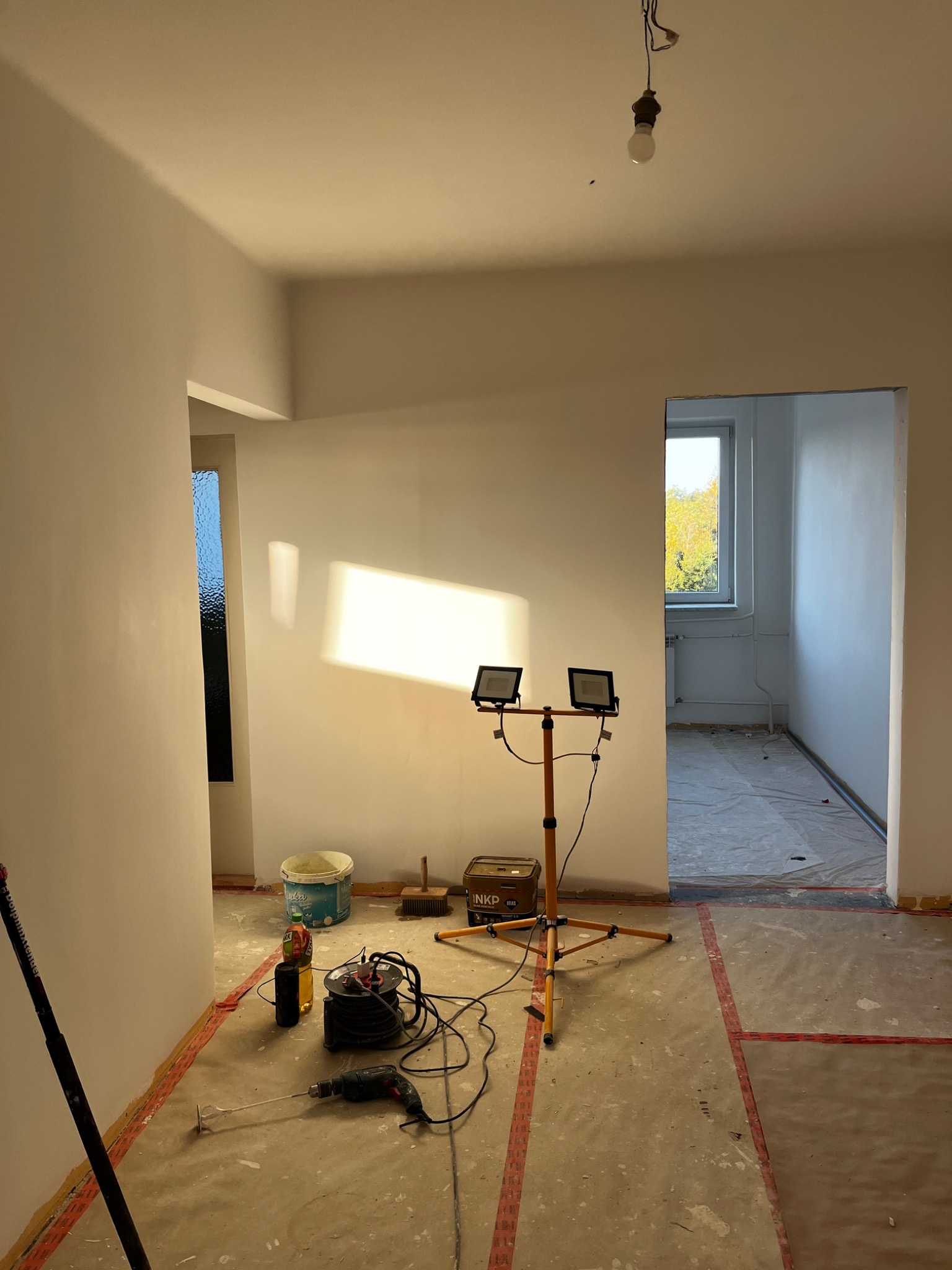 Wnętrze w trakcie malowania: białe ściany, lampa budowlana, narzędzia malarskie i zakryta podłoga. Widok na sąsiednie pomieszczenie z oknem.