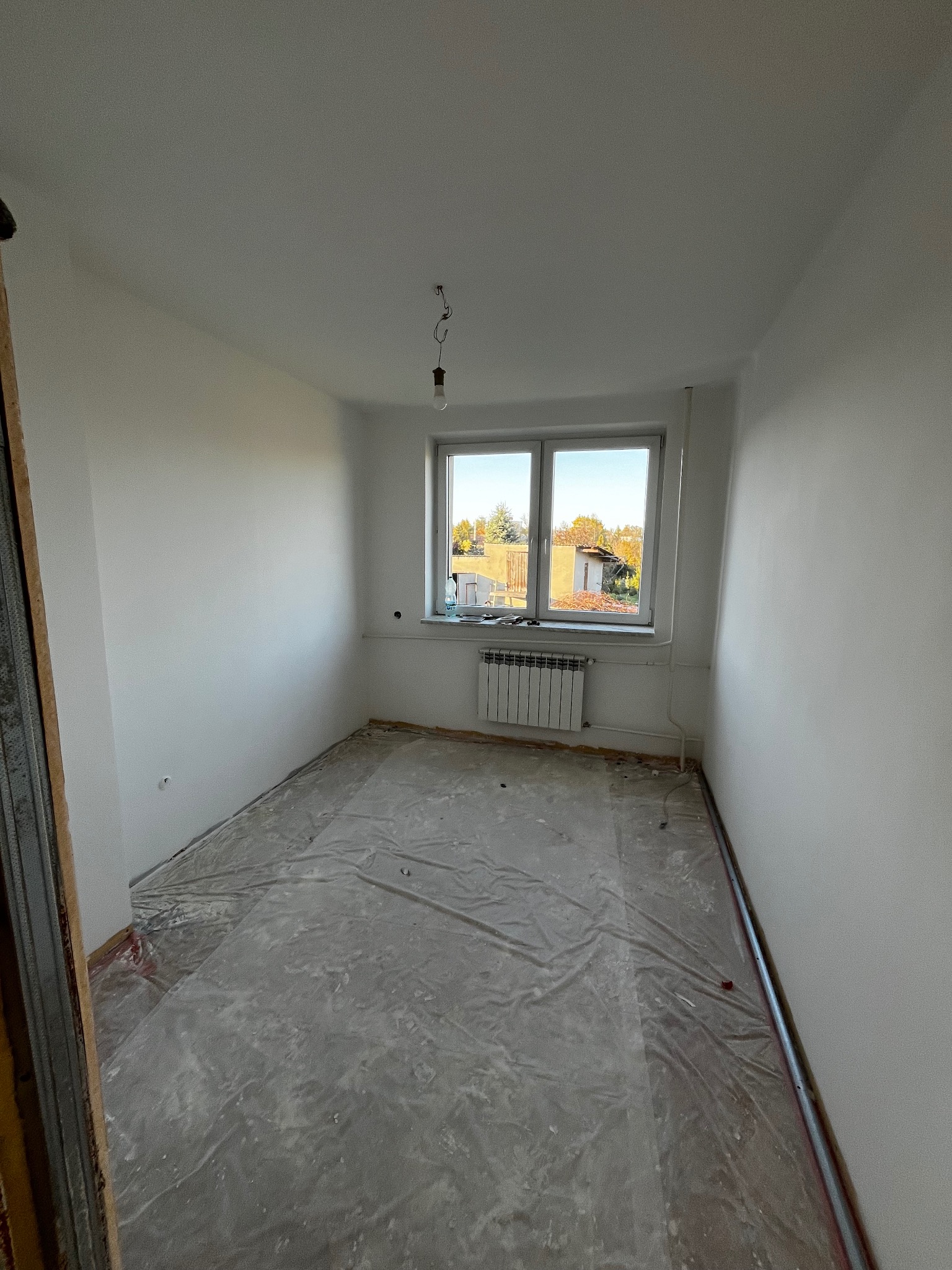 Wnętrze pokoju po malowaniu ścian na biało w Myszkowie. Podłoga zabezpieczona folią, widoczne okno i grzejnik. Ściany świeżo pomalowane, surowe wykończenie.