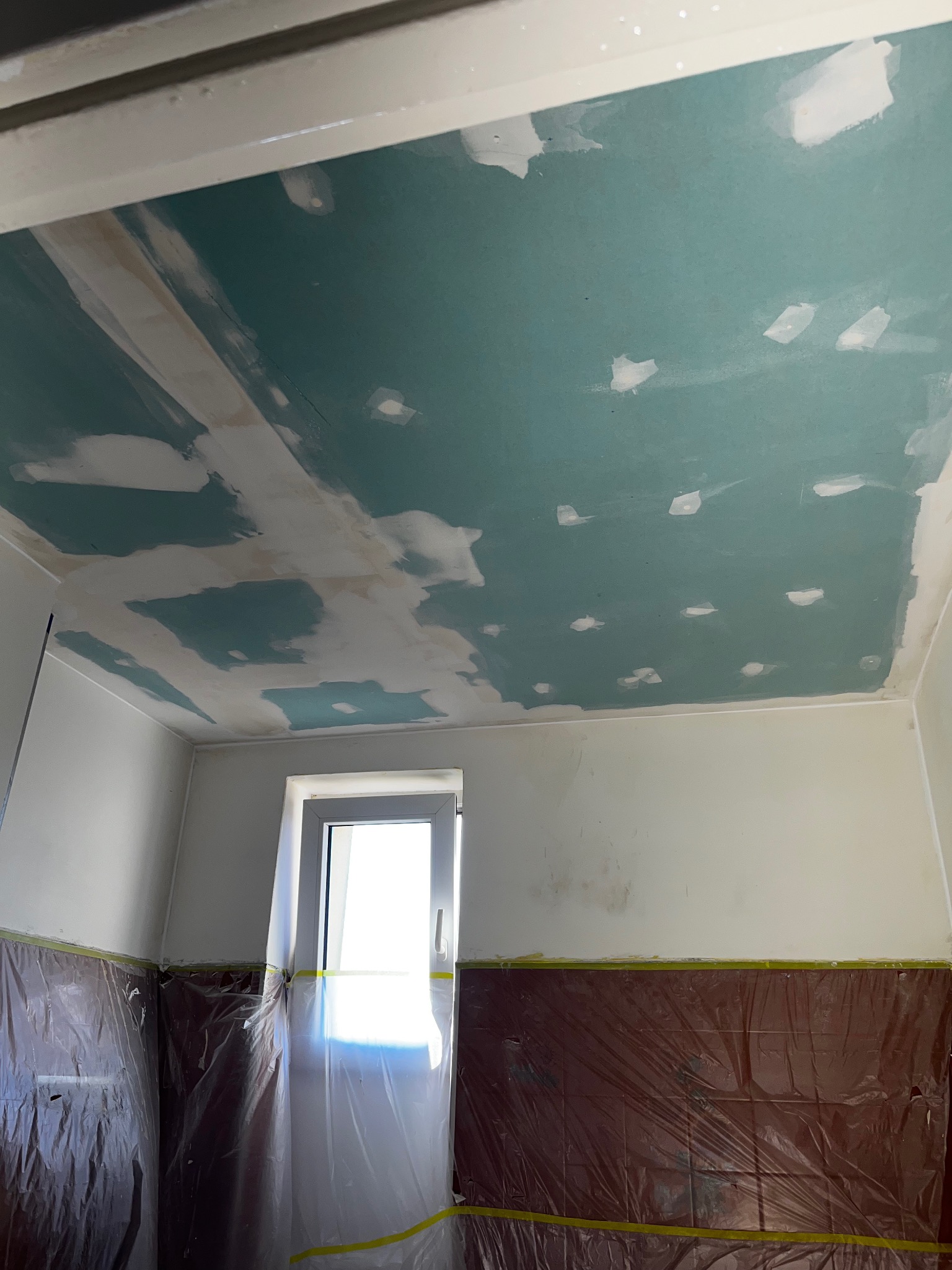 Niebieski sufit z płyt kartonowo-gipsowych w trakcie szpachlowania, zabezpieczone ściany folią malarską. Widok na okno w remontowanym pomieszczeniu.